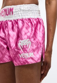 Κοντινή λήψη ενός ατόμου που φοράει ροζ σατέν αθλητικά σορτς Venum με λευκή μέση και λογότυπο, με το χέρι να ακουμπά στο πλάι.
