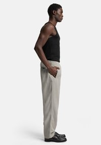Zwarte tanktop, lichtgrijze broek met een elastische tailleband, gladde textuur en zijzakken; model draagt zwarte schoenen en staat zijwaarts.