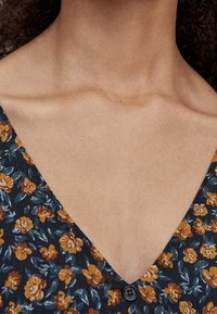 Robe florale avec un fond bleu marine orné de fleurs moutarde et blanches, décolleté en V, et un petit bouton noir au niveau de la poitrine.