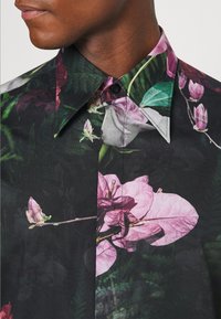 Camisa de manga corta con tela negra y vibrantes patrones florales en rosa, blanco y verde, completa con un cuello puntiagudo y detalle de botones.