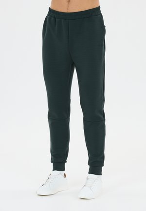 Man wearing dark green jogger pants and white sneakers standing against a plain white background.
