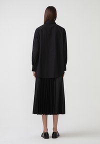 Camicia nera a maniche lunghe con colletto, indossata sopra una gonna nera a pieghe. L'outfit presenta una texture liscia e una vestibilità morbida. Scarpe nere completano il look.
