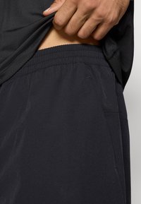 Fekete sportnadrág elasztikus derékkal, könnyű anyagból, oldalsó zsebekkel. Az anyag simának és enyhén fénylőnek tűnik.