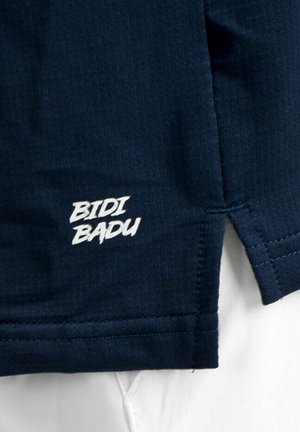 Dunkelblauer Stoff mit weiß besticktem Schriftzug "BIDI BADU" nahe einer genähten Naht und einem Saum.