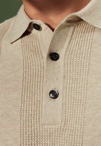 Beige pletená polo košile se strukturovaným sloupem vertikálního žebrování, dvěma černými knoflíky a klasickým límcem s plochým okrajem.