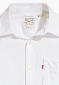 Camicia bianca Levi's con bottoni, colletto, due bottoni visibili, tasca sul petto davanti e etichetta rossa Levi's sulla cucitura della tasca.