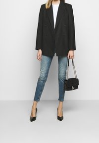 Una mujer está de pie vistiendo un blazer negro oversized, una camiseta blanca y pantalones vaqueros azules, sosteniendo un bolso negro con una correa de cadena.