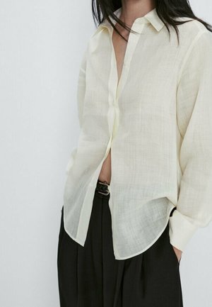 Light yellow, semi-sheer button-up shirt with a relaxed fit, long sleeves, and a curved hem. Paired with dark, loose-fitting pants.