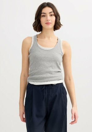 Mujer con cabello corto y castaño que lleva puestos tops sin mangas blancos y grises en capas y pantalones azul marino con cordón, de pie frente a un fondo blanco.