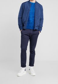 Marineblauer Bomberjacke, blauer strukturierten Pullover, dunklen Hosen und weißen Turnschuhen. Das Outfit zeichnet sich durch einen lässigen, geschichteten Look mit kontrastierenden Texturen aus.