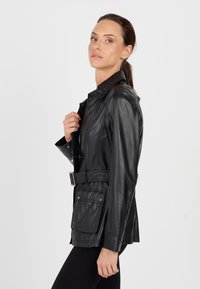 Kobieta z ciemnym kucykiem nosząca czarną skórzaną kurtkę z paskiem i czarne spodnie na jednolitym białym tle.