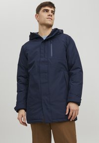 Giacca impermeabile blu navy con tasca sul petto con zip, cappuccio e tasche laterali. Presenta una texture liscia e un design leggero.