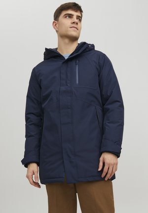 Chaqueta impermeable azul marino con un bolsillo en el pecho con cremallera, capucha y bolsillos laterales. Presenta una textura suave y un diseño ligero.