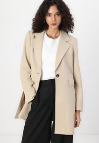 Blazer oversize beige avec des revers, fermeture à un bouton et deux poches. Porté sur un t-shirt blanc et un pantalon large noir.