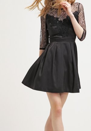 Czarna sukienka z dopasowaną górą ozdobioną koronkowymi detalami i rękawami o długości do połowy przedramienia. Rozkloszowana spódnica wykonana z gładkiego materiału.