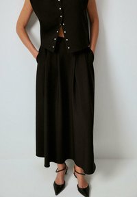 Svart ärmlös topp med tryckknappar, kombinerad med en lång, plisserad svart kjol. Bärs med svarta pumps med spetsig tå och ankelremmar.