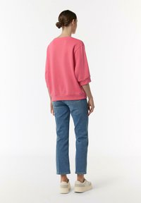 Różowy sweatshirt z opadającymi ramionami, w parze z jasnoniebieskimi dżinsami i beżowymi trampkami. Materiał wydaje się być miękki, z luźnym krojem.