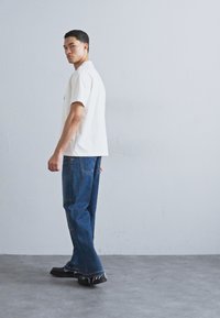 Camisa blanca de manga corta, jeans de mezclilla azul y zapatos negros. La figura está de lado contra un fondo claro y liso.