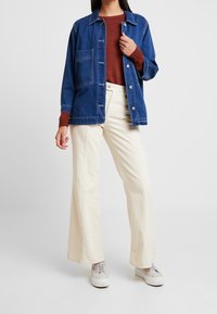 Blauwe denim jacket met grote voorzak, gedragen over een roestkleurige trui. Een paar crèmekleurige wijde broekspijpen en lichte sneakers maken de look compleet.