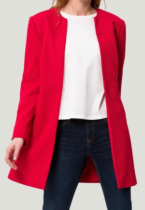 Mujer con un blazer largo rojo brillante sobre una camisa blanca y jeans azul oscuro, de pie contra un fondo claro y liso.