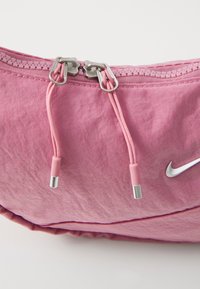 Rosa Stofftasche mit einem Reißverschluss und silbernen Metallzippverschlüssen, strukturierten Oberflächen und einem kleinen weißen Nike-Logo an der Seite.