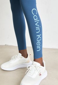 Mėlynos pėdkelnės su šviesiai mėlynu "Calvin Klein" logotipu šone, derinamos su baltomis sportinėmis bateliais, turinčiais raudonus akcentus ir tekstūruotą tinklelį.