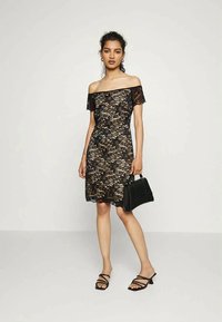 Vestido preto de renda, sem mangas, com padrões florais e um silhueta ajustada. A modelo segura uma bolsa preta; usa sandálias pretas com tiras.