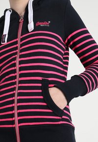Felpa nera con zip, caratterizzata da audaci strisce rosa e nere, con cerniera frontale, tasche e cordini bianchi. Logo "SuperDry" ricamato sul petto.
