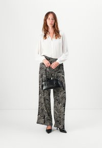 Hvit skjorte med knapp, mønstrede vide bukser i svart og krem, svart strukturert håndveske, svarte spisse sko. Minimalistisk bakgrunn.