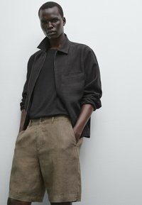 Svart linzip-jacka med en bröstficka, kombinerad med ljusbruna linshorts, båda med avslappnad passform, mot en neutral bakgrund.