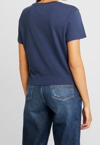 T-shirt blu navy con maniche corte, scollatura rotonda e un leggero dettaglio sulla cucitura posteriore, abbinato a jeans di denim blu scuro con effetto usato.
