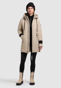 Beige gesteppte Pufferjacke mit Kapuze, Seitentaschen und schwarzen Akzenten. Kombiniert mit einem schwarzen Reißverschlussoberteil und Leggings, dazu beigen Stiefeletten.