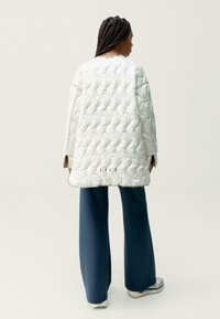 Prošívaná bílá oversized bunda s texturovaným povrchem, spárovaná s širokými modrými kalhotami, navržená v uvolněném střihu a s minimálními detaily.