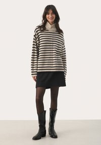 Pullover rayé chaud en beige et noir, avec col haut, coupe décontractée, porté sur une jupe noire courte et associé à des bottes noires hautes.