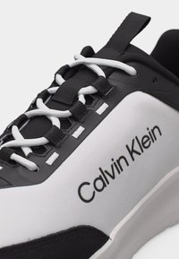 Μαύρο και άσπρο αθλητικό παπούτσι Calvin Klein με λευκά κορδόνια, μαύρες οπές και μαύρη ταινία τραβήγματος σε λευκή σόλα.