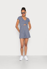 Vestido azul marinho com padrão floral, com um recorte na cintura, mangas curtas e um corte relaxado. Usado com ténis brancos e óculos de sol.