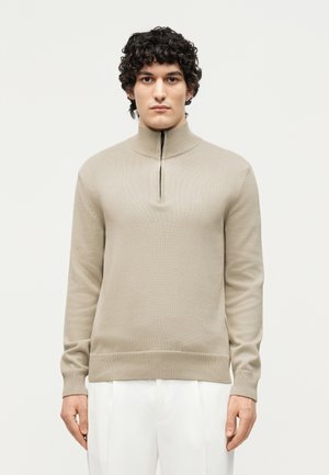 Mand med krøllet mørkt hår iført en beige strikket trøje med lynlås og hvide bukser, stående mod en ensfarvet lys baggrund.