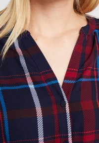 Blondynka w granatowej koszuli w kratę z czerwonymi, białymi i niebieskimi liniami, odkrywająca dekolt i górną część klatki piersiowej.