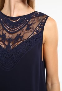 Haut sans manches bleu marine avec un col en dentelle élaborée ornée de motifs floraux et un corps en tissu lisse.