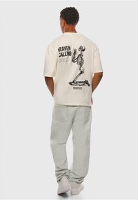 Off-white übergroßes T-Shirt mit einem bedruckten Skelett-Graphic und Text auf der Rückseite, kombiniert mit hellbeigen Hosen und weißen Sneakers.