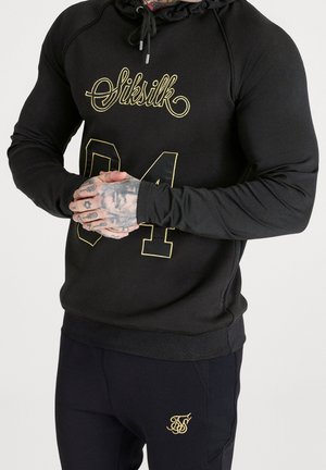 Sudadera negra con el logo dorado de "SikSilk" y el número "04" en el frente. Presenta una tela texturizada y capucha con cordón. Combinada con pantalones oscuros.