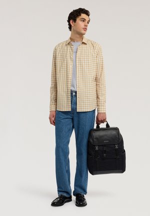 Jonge man, gekleed in een beige geruit overhemd, lichtgrijs T-shirt, blauwe spijkerbroek en zwarte schoenen, houdt een zwarte leren en stoffen rugzak aan het handvat vast.