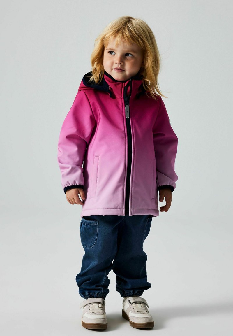 Rosa Farbverlauf Regenjacke mit einer schwarzen Kapuze, Reißverschluss vorne und elastischen Bündchen, getragen über blauen Hosen und weißen Schuhen mit brauner Sohle.
