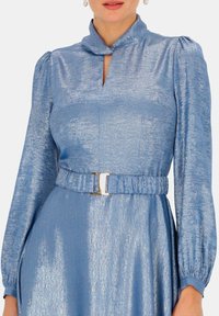 Niebieska metaliczna sukienka z wysokim kołnierzem, długimi bufkami i detalem w kształcie dziurki. Posiada dopasowany pas z srebrnym paskiem. Gładka tekstura.