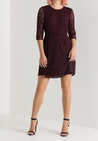 Robe en dentelle bordeaux avec des manches trois-quarts, taille cintrée et jupe évasée. Présente un décolleté rond et des accents en dentelle transparente.