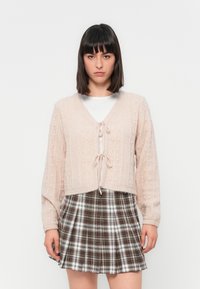 Gilet en tricot rose clair avec un décolleté en V, une fermeture à nouer et des motifs texturés; associé à une jupe plissée à carreaux marron et blanc.