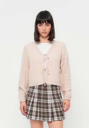 Gilet en tricot rose clair avec un décolleté en V, une fermeture à nouer et des motifs texturés; associé à une jupe plissée à carreaux marron et blanc.