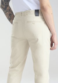 Beżowe spodnie o dopasowanym kroju, wykonane z bawełny, z dwiema tylnymi kieszeniami oraz czarną skórzaną metką przy pasku. Gładka faktura, bez widocznych wzorów.
