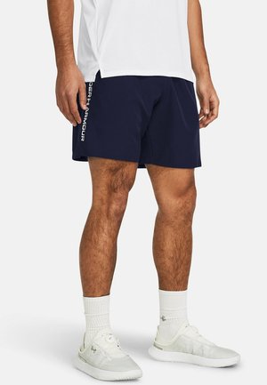 Mężczyzna w granatowych szortach z białym logo "Under Armour" na boku, białe buty sportowe, białe skarpety sportowe i biała koszulka.