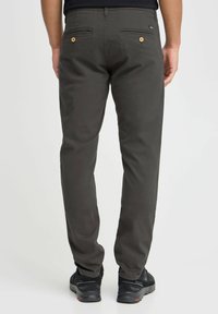 Donkergrijze broek met een rechte pasvorm, met twee achterzakken met knoopsluitingen en een gladde textuur. Draagt met zwarte schoenen.